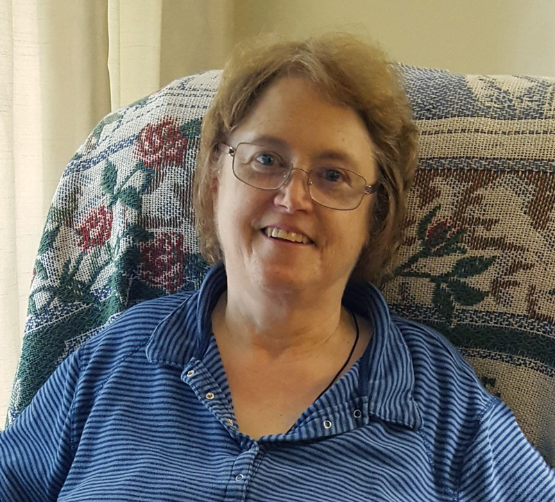 Carol smiles wearing a blue shirt and sits in a recliner chair with a throw blanket over the back 
