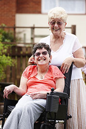 An elderly woman smiles standing behind her adult daughter in a wheelchair